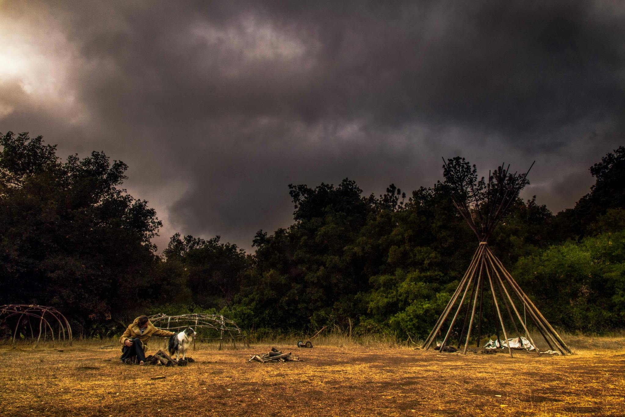 Dark sky over a wilderness campsite with a teepee and person tending to a fire, creating an adventurous mood.